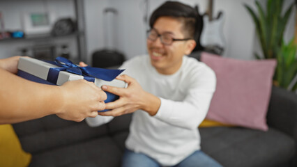 Fototapeta premium An asian man excitedly receiving a gift from another person in a cozy living room setting.