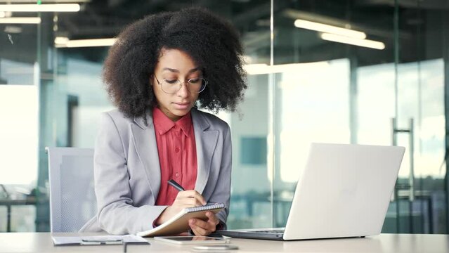 Young african american businesswoman watching video call online conference taking notes looking at laptop computer screen sitting at workplace in office. Black woman listening remote business training