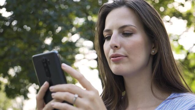 A beautiful young Caucasian woman looks at a smartphone with a smile as she sits on a bench in a park - closeup