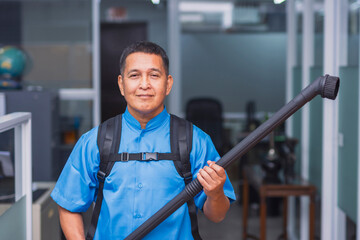 A professional janitor in blue uniform holding a round brush nozzle of a commercial backpack vacuum cleaner in an office environment. - obrazy, fototapety, plakaty