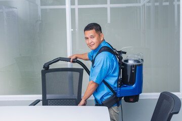A professional middle-aged janitor using a backpack vacuum cleaner to tidy up an office chair and...