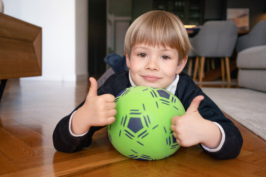 Handsome child boy holds soccer ball and gesturing thumbs up lying on floor home