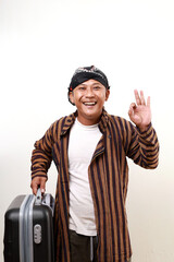 Asian adult man in javanese costume carrying suitcase while making okay hand gesture. Back to hometown concept