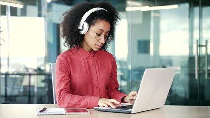 Young african american female IT specialist in headphones typing on laptop sitting at workplace in business office. Serious confident black woman developer works on computer while listening to podcast