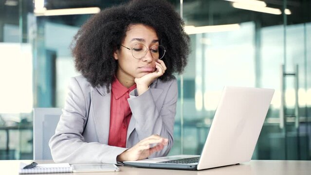 Overworked young african american businesswoman is bored sitting at workplace in office. Sleepy exhausted black woman is tired from working with a laptop. A lazy worker procrastinates, gets distracted