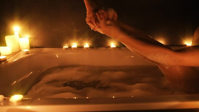 wet woman hand gently touching washing cleaning healthy skin sexy legs part of body, takes bath with soap foam girl bathes warm water white bathtub. Relax calm aroma candles light dark home spa salon