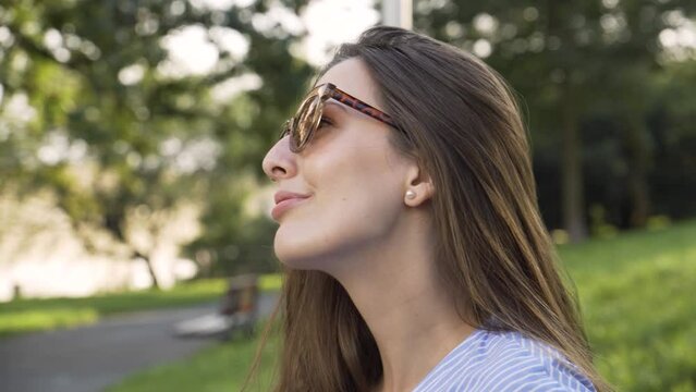 A beautiful young Caucasian woman in sunglasses looks around with a smile in a park - closeup