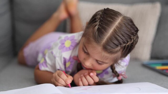 Adorable little girl with beautiful braid hairstyle lying on sofa at home and drawing picture for mom
