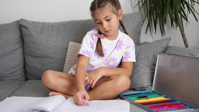 Cute little girl sitting on sofa in living room and draws by colored pencils. Home mood. Braid hairstyle