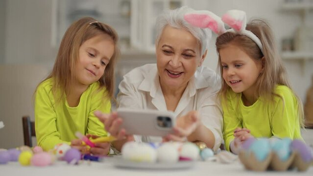 Easter grandmothers with granddaughters. Smiling mature grandmother with twins grandchildren watching cartoons on phone in rabbit bunny ears, talking, celebrating close up. Easter holiday concept.