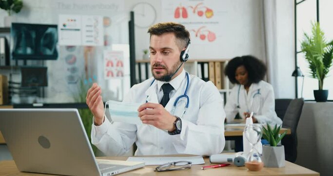 Man doctor in wireless headset using laptop telling to online audience about the need to wear protective mask
