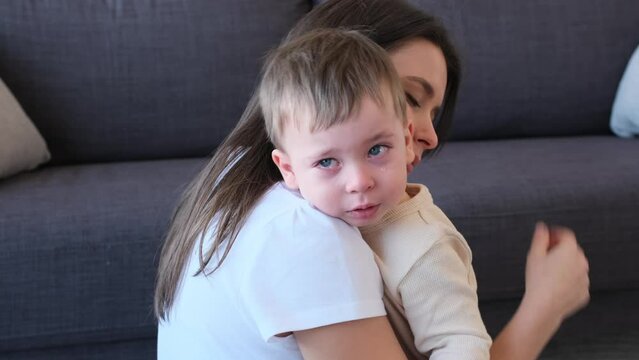 Caring young Caucasian mother embracing her crying little son at home living room. Displeased and annoyed toddler, with tears on face.