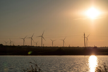 Windmills with rotor blades generating green energy on riverbank at sunset light. Production of...