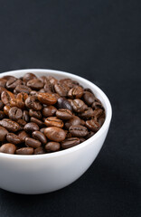 Part of a white bowl with roasted Arabica coffee beans close-up on a dark background