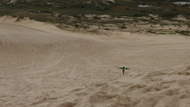 Boy sandboarding at dunes walks up sand hill carrying board - wide shot
