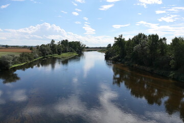 A horizontal view of a serene river in France during the summer. The water is low, and trees line the riverbanks, their vibrant green foliage reflected in the calm waters.