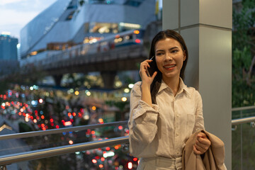 Businesswoman standing on the sky train station talking, speaking, chatting on brand name smart phone while waiting for the train after work overtime. Bangkok, Thailand.