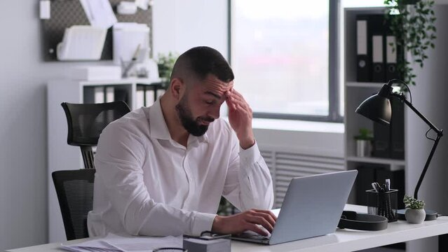 Unhappy and weary Caucasian male office worker, sitting at desk with laptop, working overtime in office. Emotional stress and tiredness concept.