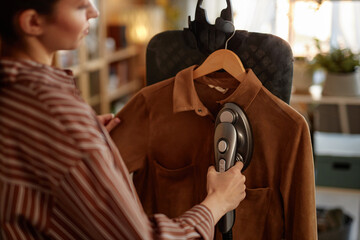 Close up of young woman using garment steamer on vertical ironing board and steaming suede jacket...