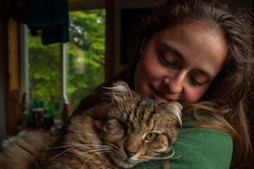 A woman holds a long-haired Tabby Cat in her arms inside by a window