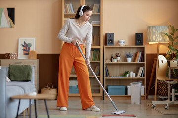 Full length portrait of young woman wearing headphones and mopping floors at home while enjoying...