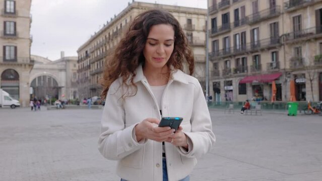 Cheerful curly beautiful young woman in casual clothes using mobile phone while walking at city street.Technology and urban lifestyle concept