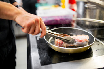 A person cooks steaks using tongs on a stovetop pan in a well-lit kitchen. The electric stovetop has visible heating elements, and various kitchen items are in the background