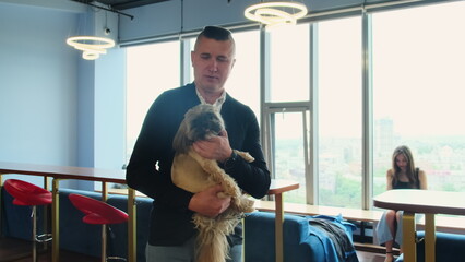 Young businessman in casual clothes holds calm Shih Tzu in arms. Man stands in office accompanied...