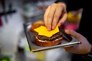 hand places a bright yellow slice of cheddar cheese onto a grilled burger patty, creating a...