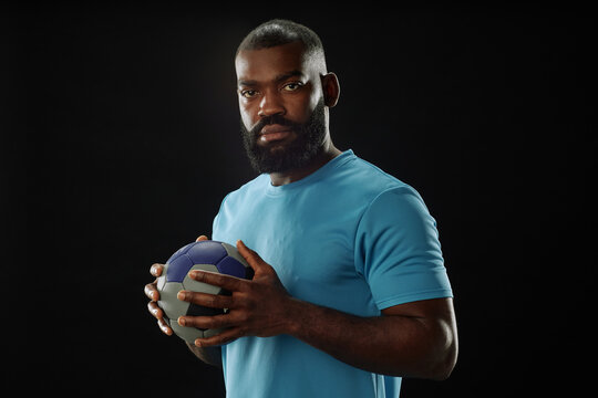 Minimal waist up portrait of African American football player holding ball and looking at camera against black background in studio copy space