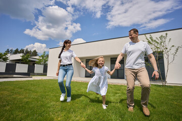 Pleased family holding hands on green yard near modern townhouse in summer day