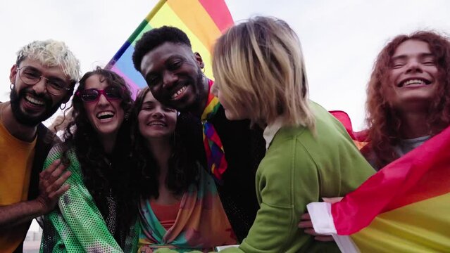 Multiracial group of young people celebrating Gay Pride Festival Day with rainbow flags. LGBT community concept with laughing men and women hugging looking at camera outdoors.
