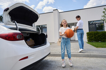 Little girl with basketball ball jumping near her mother carrying cardboard box