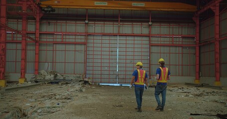 Fototapeta premium Two construction workers in yellow hard hats and reflective vests discussing stand amidst a large industrial site, examine a building site with structural steelwork in the background
