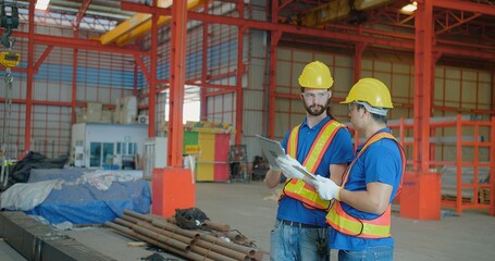 Two construction workers in reflective vests and hard hats consulting over a clipboard in an industrial warehouse, symbolizing teamwork and project planning