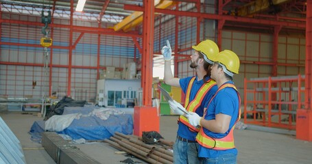 Two construction workers in reflective vests and hard hats consulting over a clipboard in an industrial warehouse, symbolizing teamwork and project planning