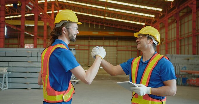 Two smiling construction workers in safety gear high-five, celebrating teamwork and successful collaboration at a well-lit industrial site.