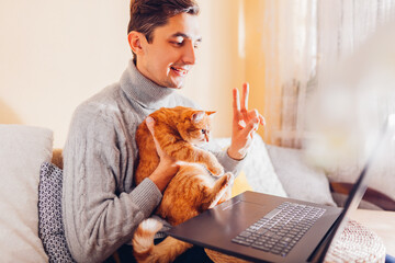 Man facetiming from home holding pet using computer. Ginger cat looking at laptop screen during video chat.