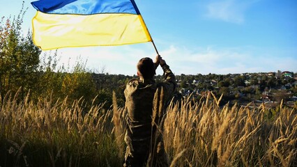 Male military in uniform waving flag of Ukraine at countryside. Young soldier of ukrainian army lifting blue-yellow banner as symbol of victory against russian aggression. Invasion resistance concept. - 756617615