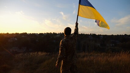 Male soldier stands with raised flag of Ukraine against background of beautiful sunset. Male ukrainian army soldier with a lifted blue-yellow banner in honor of the victory against russian aggression - 756617880