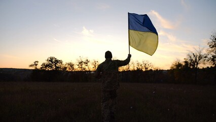 Soldier of ukrainian army running with raised blue-yellow banner on field at dusk. Young male military in uniform jogging with flag of Ukraine at meadow. Victory against russian aggression concept - 756618067