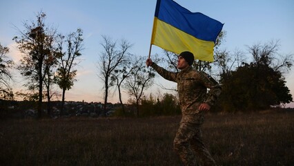 Soldier of ukrainian army running with raised blue-yellow banner on field at dusk. Young male military in uniform jogging with flag of Ukraine at meadow. Victory against russian aggression concept - 756618206