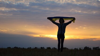 Happy lady walking on barley meadow with a raised over head flag of Ukraine. Ukrainian woman with a lifted blue-yellow banner on a beautiful sunset at background. End of war concept. - 756618454