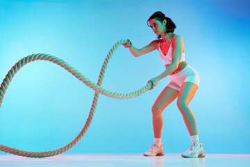 Sportive, slim young woman with strong muscular body training with ropes against blue studio background in neon light. Concept of sport, health and body care, fitness app, exercises templates