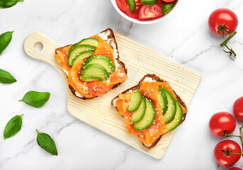 Toasts or open sandwiches with avocado, smoked salmon, sesame seeds and cream cheese on a wooden cutting board, with tomatoes and basil leaves lying around. Homemade food concept. Flat lay, top view