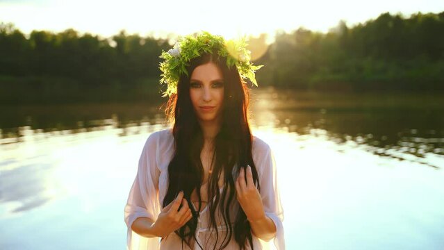 fantasy wet woman stands in lake, hand stroking touching long dark hair water herbal wreath flowers. Sexy girl fashion model beauty face looking at camera enjoy summer nature sun light sunset tree 
