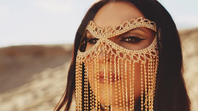 Close-up portrait young arabian woman brown eyes oriental golden makeup look camera. Gold metal chain golden mask hides face. black hair fly in wind sexy fashion model girl luxury glamour style art