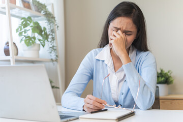 Exhausted, asian young business woman working at office desk, took off eye glasses, massage rubbing nose from dry eyes, suffer on work hard, using laptop computer at office home, problem overtime job.