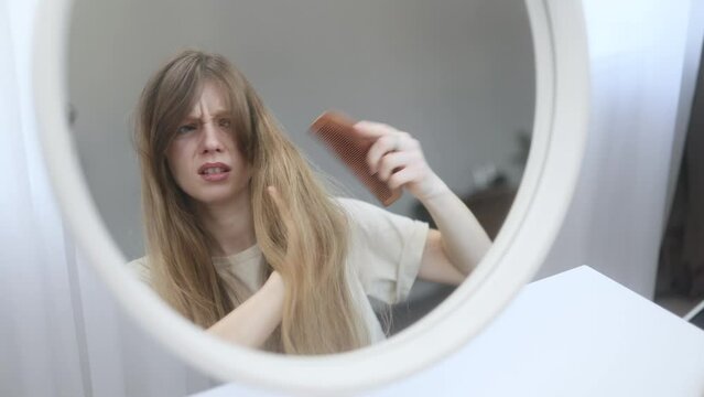 Portrait of nervous caucasian young woman having difficulty brushing her tangled hair dryness split ends problem by wooden brash while looking at mirror reflection at home