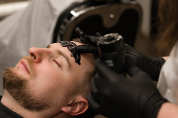 Barber applies a black moisturizing mask to a man's face while shaving. A dermatologist applies a...